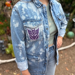 May include: A light blue acid-wash denim jacket featuring a purple and white Decepticon logo patch on the chest pocket. The jacket has a classic collar, long sleeves with folded cuffs, and metal button closures down the front and on the pockets.