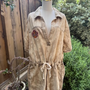 May include: A tan-colored romper with a collared neckline and short sleeves. The romper features a drawstring waist, a small patch on the chest, and a bleached pattern. The garment is displayed on a mannequin outdoors.