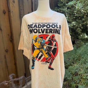 May include: A light peach-colored t-shirt featuring a graphic of Deadpool and Wolverine in action poses. The shirt has the text "DID WE JUST BECOME BEST BUBS?!" and "DEADPOOL & WOLVERINE" printed on the front. The Marvel Studios logo is also visible.