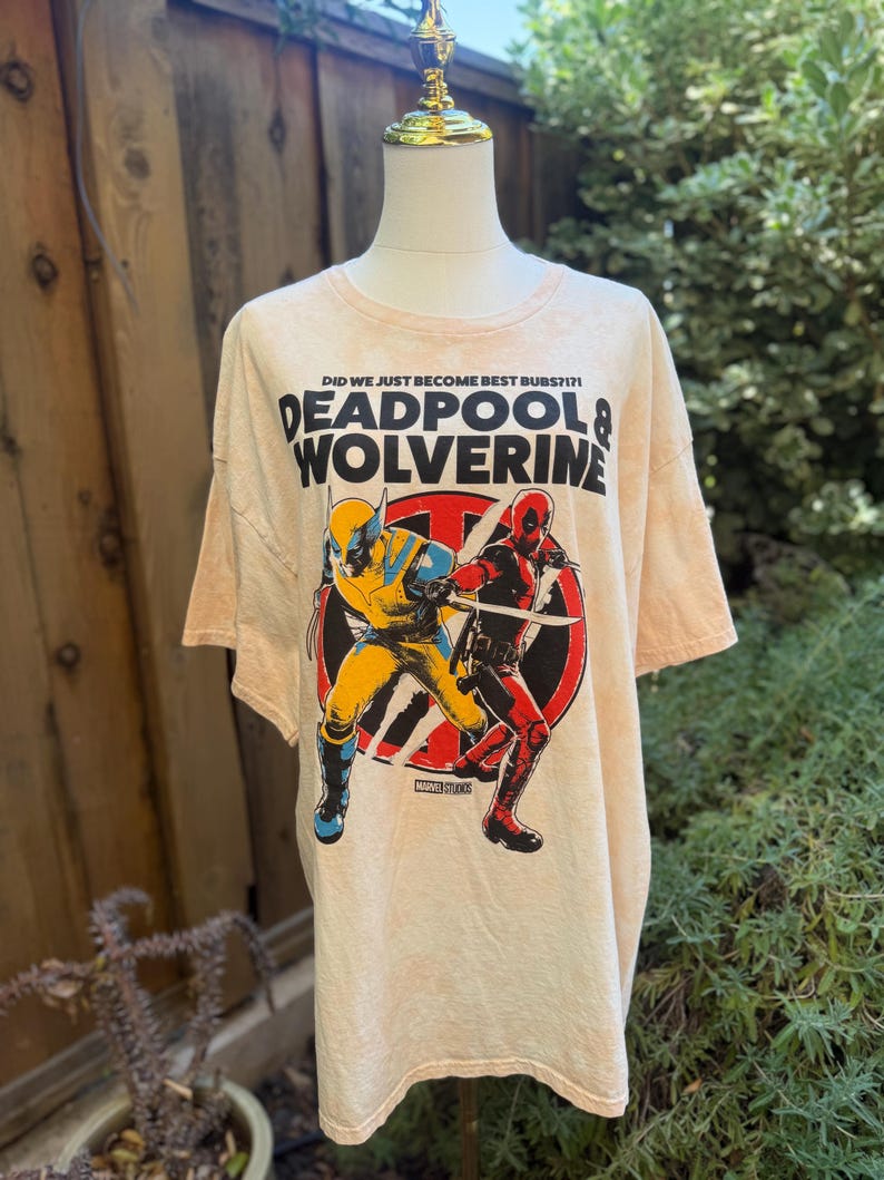 May include: A light peach-colored t-shirt featuring a graphic of Deadpool and Wolverine in action poses. The shirt has the text "DID WE JUST BECOME BEST BUBS?!" and "DEADPOOL & WOLVERINE" printed on the front. The Marvel Studios logo is also visible.
