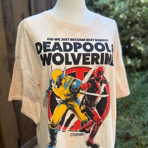 May include: A light beige t-shirt featuring a graphic of Deadpool and Wolverine in action poses. The text "DEADPOOL & WOLVERINE" is printed in large, bold letters above the characters. The shirt also includes the text "DID WE JUST BECOME BEST BUBS?!" and the Marvel Studios logo.