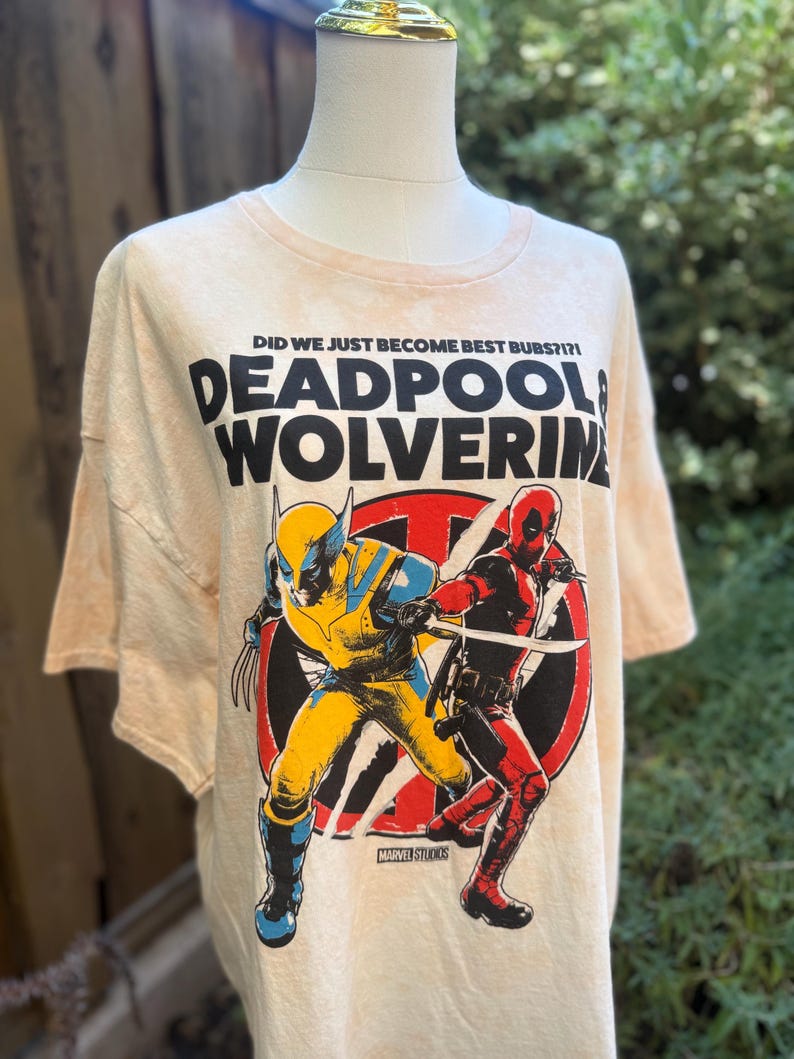May include: A light beige t-shirt featuring a graphic of Deadpool and Wolverine in action poses. The text "DEADPOOL & WOLVERINE" is printed in large, bold letters above the characters. The shirt also includes the text "DID WE JUST BECOME BEST BUBS?!" and the Marvel Studios logo.