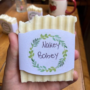 Puede incluir: Una barra de jabón artesanal con superficie texturizada, sostenida en una mano. El jabón tiene una etiqueta blanca con las palabras "Nakey Babey" dentro de una corona de hojas verdes. Otras barras de jabón son visibles en el fondo.