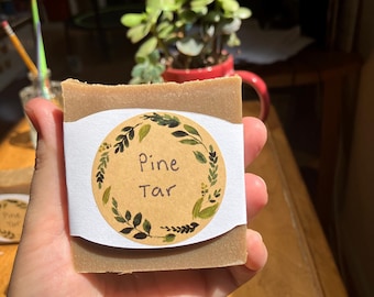 Peut inclure: Une barre de savon brune avec une étiquette blanche qui dit "Pine Tar" dans un cercle entouré de feuilles vertes.