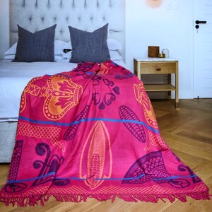 May include: A bright pink blanket featuring a detailed pattern in orange, purple, and blue. The blanket is draped over a bed, displaying its fringed edge and intricate designs. The bed has grey pillows and a white headboard.