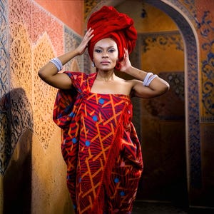 May include: A woman wearing a red head wrap and a matching patterned wrap. The wrap has a geometric design in orange and blue on a red background. The text "HIMBA HERITAGE THROW" is visible in the image.