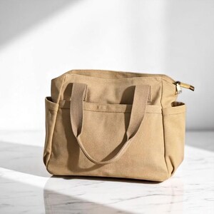 May include: A tan canvas tote bag with two handles and side pockets. The bag has a zipper closure and a rectangular shape. The handles are a lighter shade of tan. The bag is sitting on a white surface.