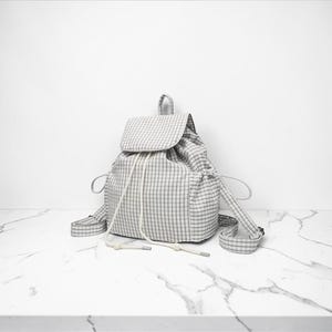May include: A grey and white checked backpack with a flap and drawstring closure. It features adjustable straps and a small top handle. The bag is made of soft fabric and is placed on a white marble surface.