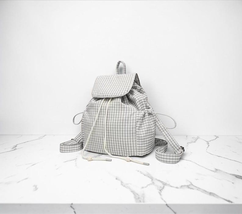 May include: A grey and white checked backpack with a flap and drawstring closure. It features adjustable straps and a small top handle. The bag is made of soft fabric and is placed on a white marble surface.