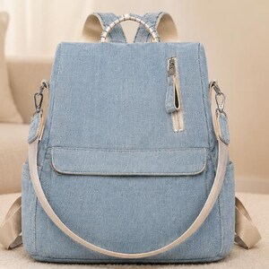 Puede incluir: Una mochila de mezclilla azul claro con un bolsillo vertical con cremallera en la parte delantera, un bolsillo horizontal con solapa y correas beige para los hombros. La bolsa incluye herrajes de metal plateado, un asa superior y bolsillos laterales.