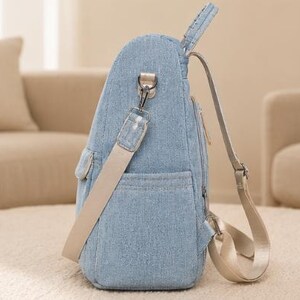 Puede incluir: Una mochila de mezclilla azul claro con un bolsillo lateral, una correa de hombro beige desmontable con un clip de metal plateado y correas de mochila beige ajustables. El bolso tiene un cierre de cremallera vertical y un perfil estructurado.
