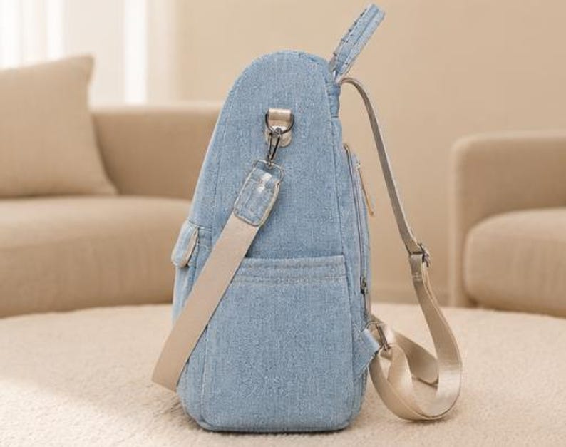 Puede incluir: Una mochila de mezclilla azul claro con un bolsillo lateral, una correa de hombro beige desmontable con un clip de metal plateado y correas de mochila beige ajustables. El bolso tiene un cierre de cremallera vertical y un perfil estructurado.