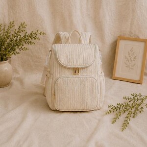 May include: A cream-coloured quilted backpack featuring a vertical ribbed texture, a top handle, adjustable shoulder straps, and a front zippered pocket. The bag includes gold-toned metal zips and side pockets, showcasing a structured design.