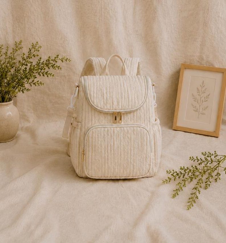 May include: A cream-coloured quilted backpack featuring a vertical ribbed texture, a top handle, adjustable shoulder straps, and a front zippered pocket. The bag includes gold-toned metal zips and side pockets, showcasing a structured design.