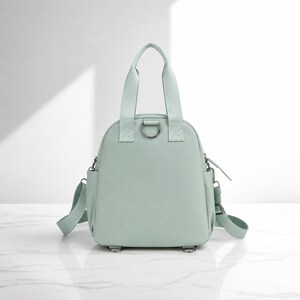 May include: A light mint green backpack with two top handles, a central metal D-ring, side pockets, and adjustable shoulder straps. The bag features a smooth, solid-coloured exterior with matching fabric straps and silver-toned metal hardware components.