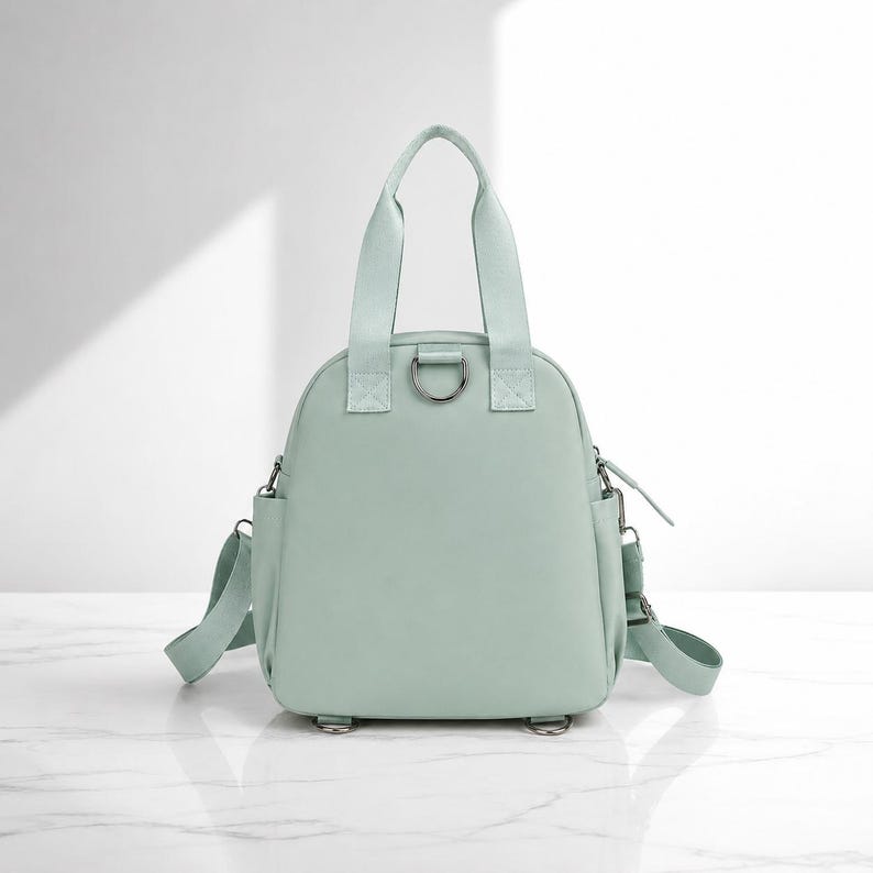 May include: A light mint green backpack with two top handles, a central metal D-ring, side pockets, and adjustable shoulder straps. The bag features a smooth, solid-coloured exterior with matching fabric straps and silver-toned metal hardware components.