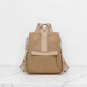 K&ouml;nnte beinhalten: Ein beigefarbener Rucksack aus Kord mit einem vertikalen Mittelstreifen, einer horizontalen Vordertasche mit geflochtenem Rei&szlig;verschlussanh&auml;nger, zwei Seitentaschen und verstellbaren Schultergurten. Die Tasche besteht aus strukturiertem Stoff.