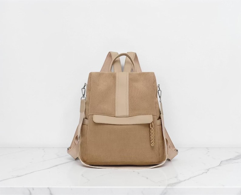K&ouml;nnte beinhalten: Ein beigefarbener Rucksack aus Kord mit einem vertikalen Mittelstreifen, einer horizontalen Vordertasche mit geflochtenem Rei&szlig;verschlussanh&auml;nger, zwei Seitentaschen und verstellbaren Schultergurten. Die Tasche besteht aus strukturiertem Stoff.