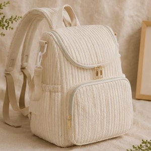 May include: A cream-coloured corduroy backpack featuring vertical ribbed texture, a top handle, adjustable shoulder straps, and a front zipped pocket. The bag includes gold-toned metal zips and side pockets, providing a structured design.