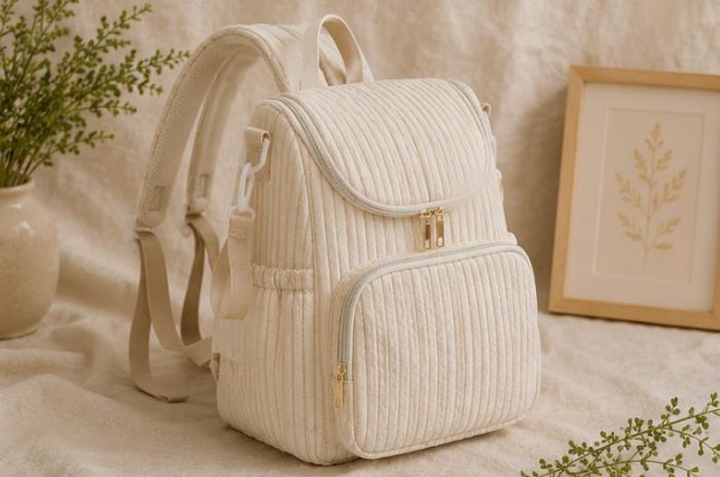 May include: A cream-coloured corduroy backpack featuring vertical ribbed texture, a top handle, adjustable shoulder straps, and a front zipped pocket. The bag includes gold-toned metal zips and side pockets, providing a structured design.