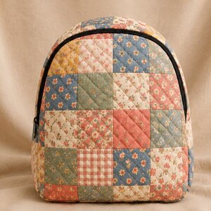 K&ouml;nnte beinhalten: Ein gesteppter Rucksack mit einem Patchwork-Design aus verschiedenen Blumen- und Vichy-Karomustern in gedeckten Blau-, Rosa-, Gr&uuml;n- und Gelbt&ouml;nen. Die Tasche hat eine abgerundete Oberseite, einen schwarzen Rei&szlig;verschluss und eine rautenf&ouml;rmige Steppung.