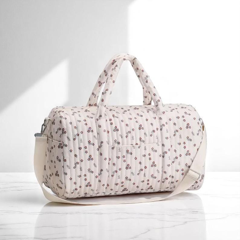 May include: A quilted duffel bag in a light cream color featuring a repeating pattern of small dark floral sprigs. The bag includes two top handles, a rectangular front pocket, and a wide, adjustable off-white shoulder strap attached at the sides.
