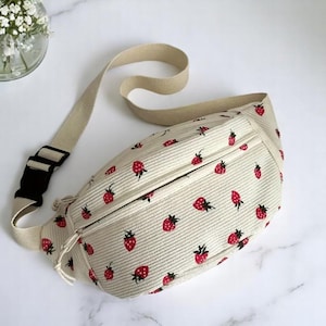 May include: Cream-colored fanny pack with a repeating pattern of red strawberries. The bag has a beige adjustable strap and a black buckle. The fanny pack is made of a corduroy-like material.