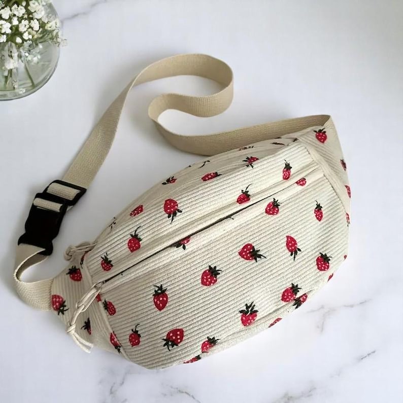 May include: Cream-colored fanny pack with a repeating pattern of red strawberries. The bag has a beige adjustable strap and a black buckle. The fanny pack is made of a corduroy-like material.