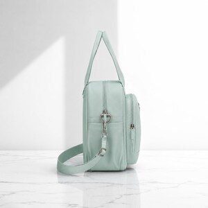May include: A light mint green backpack with a vertical rectangular shape, featuring a top handle, a detachable shoulder strap with silver metal hardware, a main zippered compartment, and a smaller front zippered pocket with a matching pull tab.