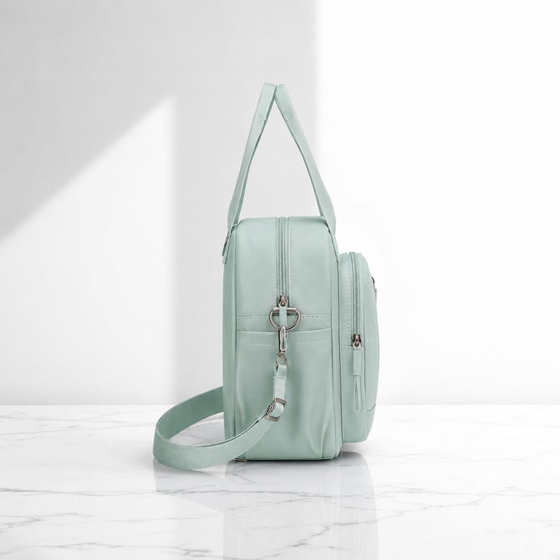 May include: A light mint green backpack with a vertical rectangular shape, featuring a top handle, a detachable shoulder strap with silver metal hardware, a main zippered compartment, and a smaller front zippered pocket with a matching pull tab.