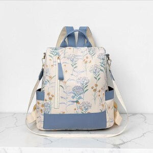 May include: A cream-coloured floral backpack with blue detailing. The bag features a pattern of blue and yellow flowers. It has a blue base, straps, and handle, with a zipped front pocket and side pockets. The bag is suitable for everyday use.