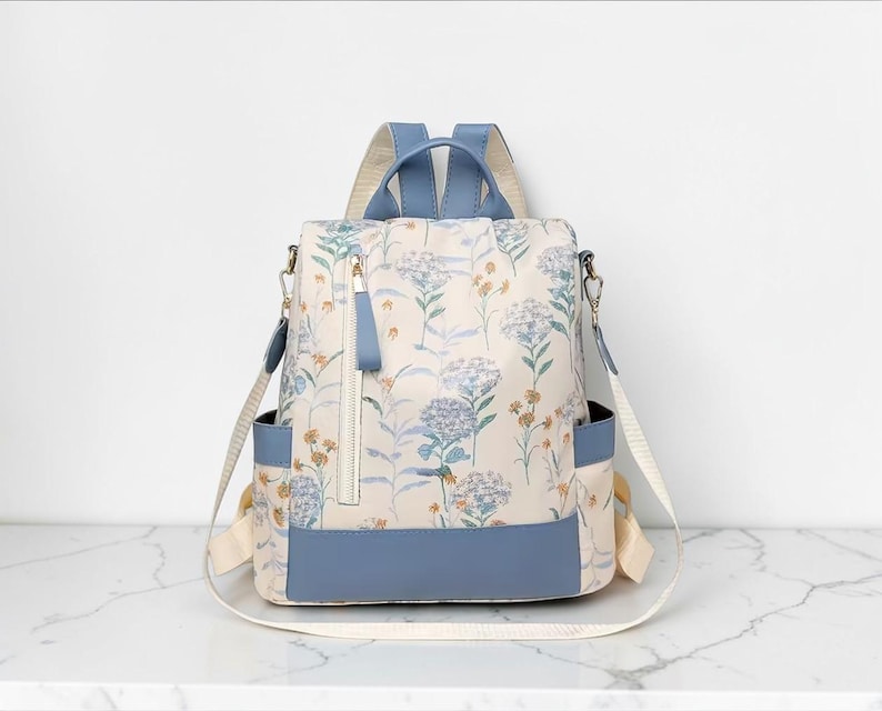May include: A cream-coloured floral backpack with blue detailing. The bag features a pattern of blue and yellow flowers. It has a blue base, straps, and handle, with a zipped front pocket and side pockets. The bag is suitable for everyday use.