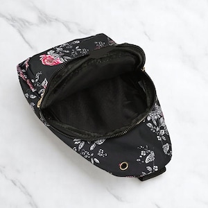 May include: A black crossbody bag with a floral print featuring white and pink flowers. The bag has a zipper closure and a gold metal buckle.