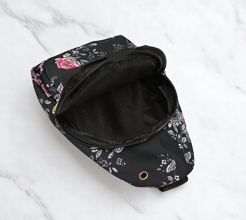 May include: A black crossbody bag with a floral print featuring white and pink flowers. The bag has a zipper closure and a gold metal buckle.
