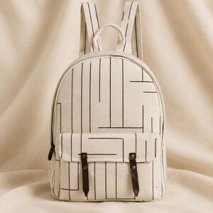 May include: A cream-colored canvas backpack featuring a black linear geometric pattern. The bag includes a front pocket with a flap secured by two dark brown leather straps with brass-colored metal rivets, a top handle, and two shoulder straps.