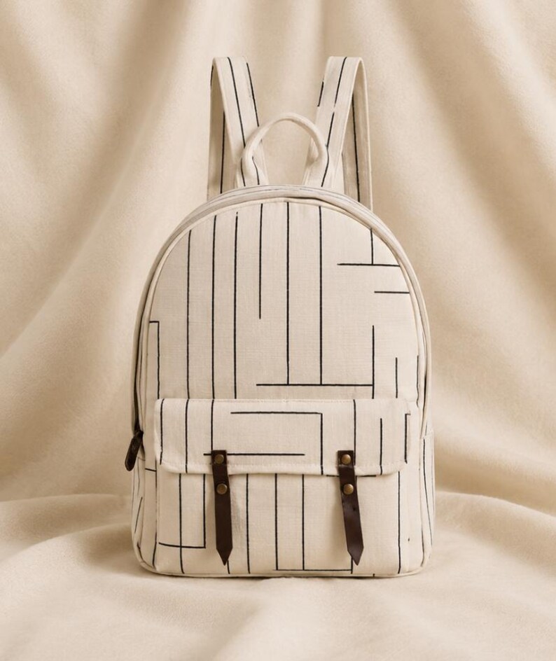 May include: A cream-colored canvas backpack featuring a black linear geometric pattern. The bag includes a front pocket with a flap secured by two dark brown leather straps with brass-colored metal rivets, a top handle, and two shoulder straps.