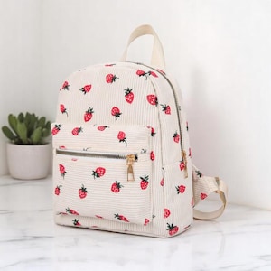 May include: A white corduroy backpack with a strawberry print. The backpack has a zippered front pocket and adjustable straps.