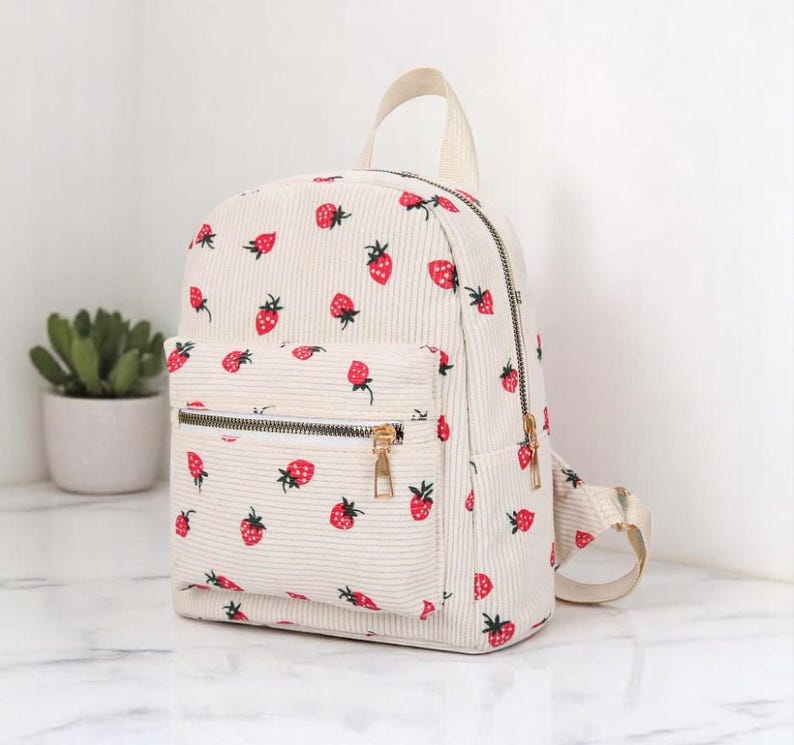 May include: A white corduroy backpack with a strawberry print. The backpack has a zippered front pocket and adjustable straps.