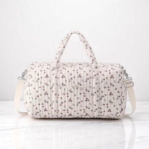 May include: A quilted duffel bag in a light cream color featuring a repeating pattern of small dark red and green floral sprigs. The bag includes two top handles, a detachable shoulder strap with silver metal hardware, and a front pocket.