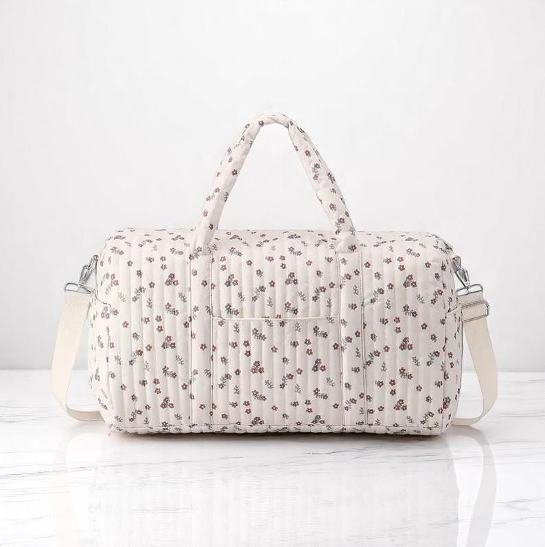 May include: A quilted duffel bag in a light cream color featuring a repeating pattern of small dark red and green floral sprigs. The bag includes two top handles, a detachable shoulder strap with silver metal hardware, and a front pocket.