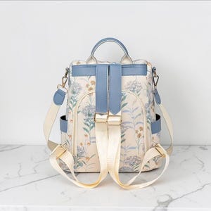 May include: A floral print backpack with a light blue top handle, straps, and accents. The backpack has a cream-coloured background with a delicate floral pattern in shades of blue and green. It features adjustable straps and a secure closure.