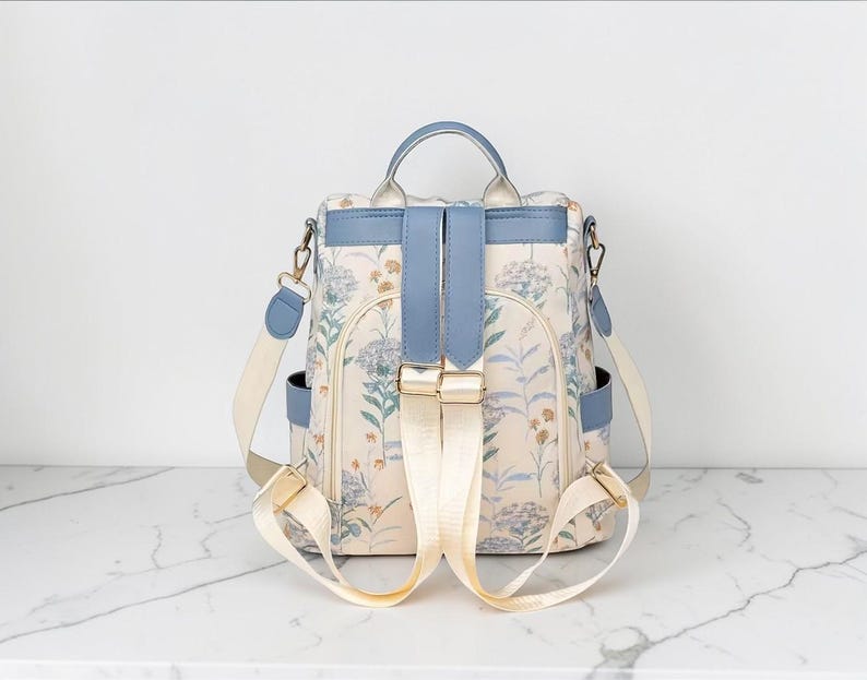 May include: A floral print backpack with a light blue top handle, straps, and accents. The backpack has a cream-coloured background with a delicate floral pattern in shades of blue and green. It features adjustable straps and a secure closure.