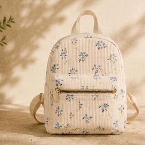 K&ouml;nnte beinhalten: Ein kleiner cremefarbener Rucksack aus Kord mit einem zarten blauen Blumenmuster. Die Tasche verf&uuml;gt &uuml;ber eine Fronttasche mit Rei&szlig;verschluss und goldfarbenen Beschl&auml;gen, einen Tragegriff oben sowie verstellbare Schultergurte im kompakten Design.