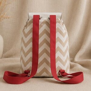 May include: A backpack featuring a beige and white chevron pattern on the main body. The bag has a solid white top flap and two wide, bright red adjustable shoulder straps attached to the back. The design includes metal hardware connectors.