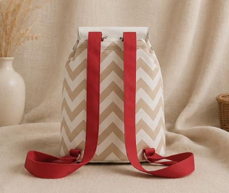 May include: A backpack featuring a beige and white chevron pattern on the main body. The bag has a solid white top flap and two wide, bright red adjustable shoulder straps attached to the back. The design includes metal hardware connectors.