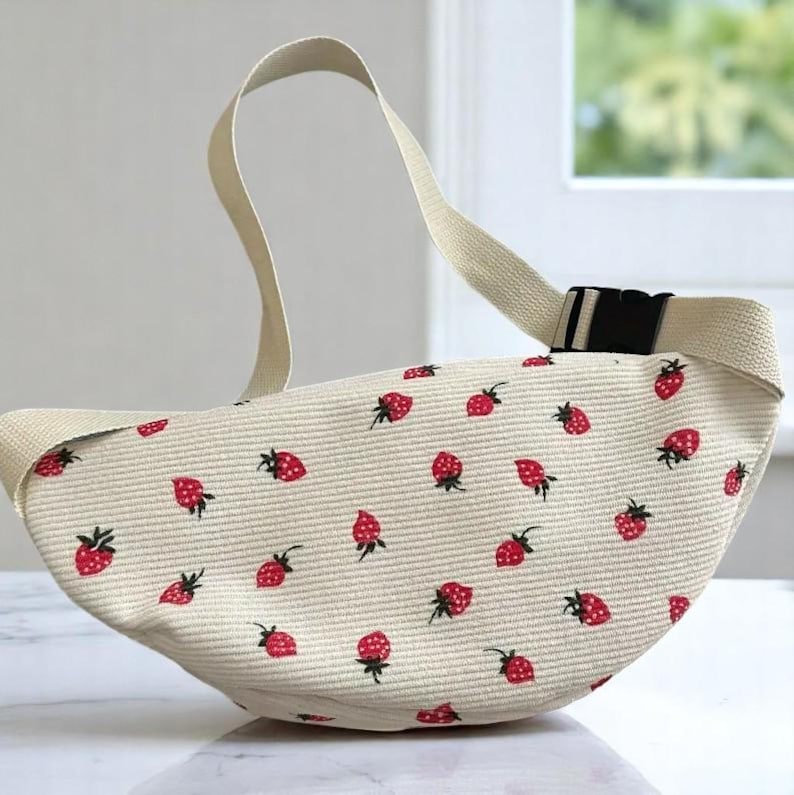 May include: Cream-colored fanny pack with a repeating pattern of red strawberries. The bag has a cream-colored strap with a black buckle. The bag is made of a textured fabric.