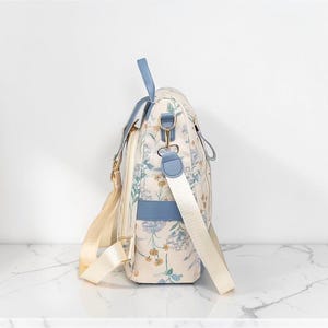 May include: A cream-coloured bag with a floral print and blue accents. The bag has a shoulder strap and a top handle. The floral pattern includes blue, yellow, and green flowers. The bag has gold-coloured hardware and a cream-coloured strap.