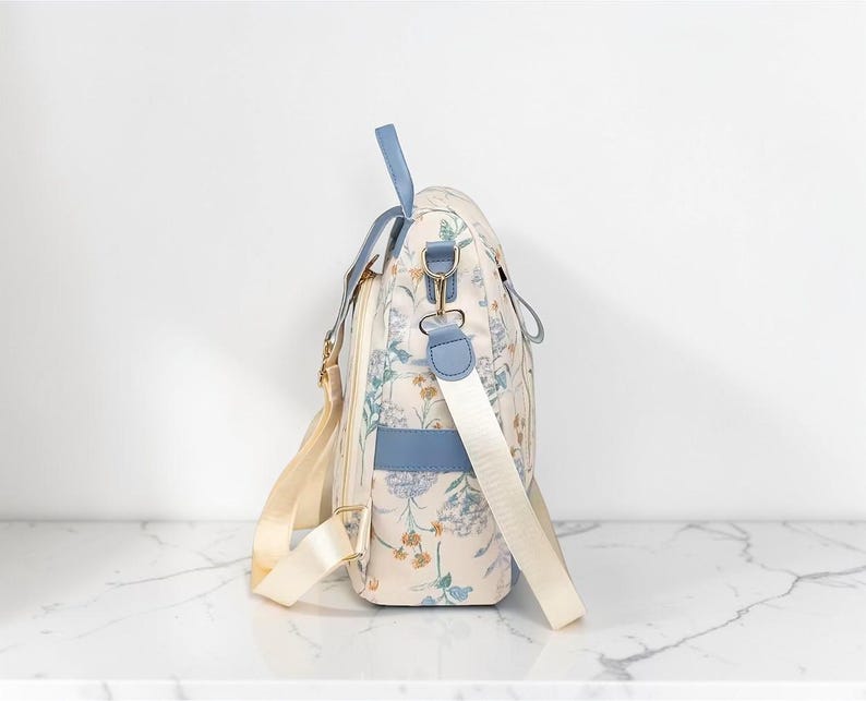 May include: A cream-coloured bag with a floral print and blue accents. The bag has a shoulder strap and a top handle. The floral pattern includes blue, yellow, and green flowers. The bag has gold-coloured hardware and a cream-coloured strap.