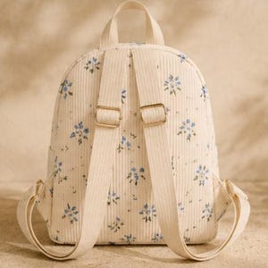 K&ouml;nnte beinhalten: Ein cremefarbener Rucksack aus Cord mit einem sich wiederholenden Muster aus kleinen blauen Blumen und gr&uuml;nen Bl&auml;ttern. Der Rucksack verf&uuml;gt &uuml;ber zwei verstellbare Schultergurte, einen Tragegriff und goldfarbene Metallbeschl&auml;ge an den Schnallen.