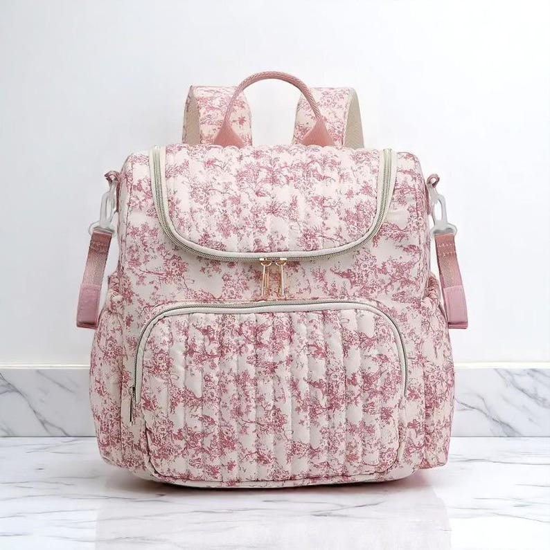 Puede incluir: Mochila acolchada con estampado floral en rosa y blanco. Cuenta con asa superior, correas ajustables y bolsillo frontal. Las cremalleras y herrajes son de color dorado.